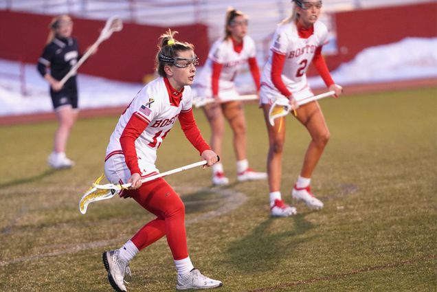 Action photo of Emma Pfaff, a women lacrosse player who is running down the field with her lacrosse stick and goggles on. Two other female BU lacrosse players can be seen in the background, on field.