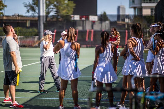 Women's tennis players stand in the shade and listen to their coach talk at BU