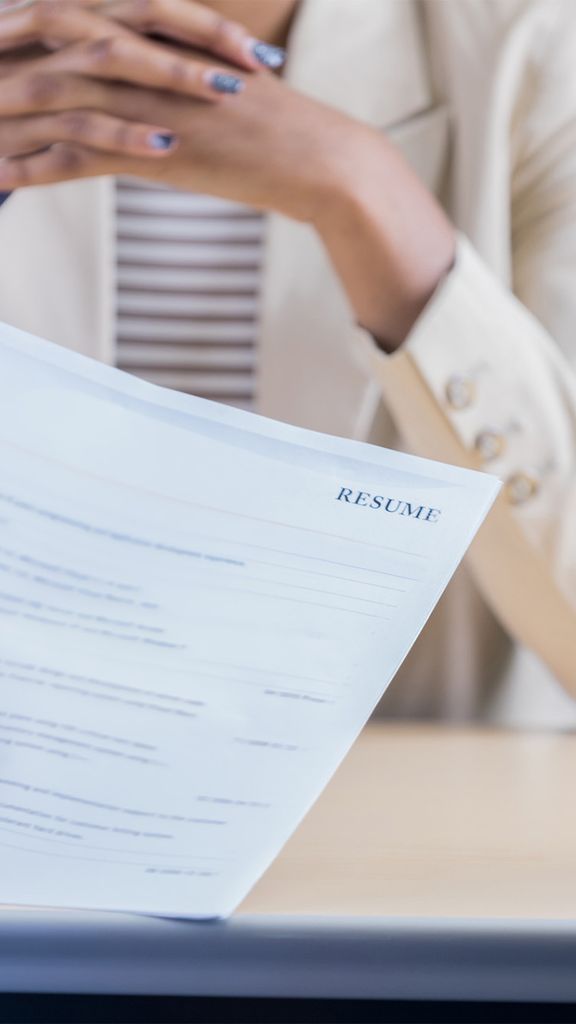Unrecognizable businessperson conducts a job interview with an unrecognizable female candidate. One hand is holding the woman's resume. The woman is sitting across the desk from them. Photo via iStock / SDI Productions