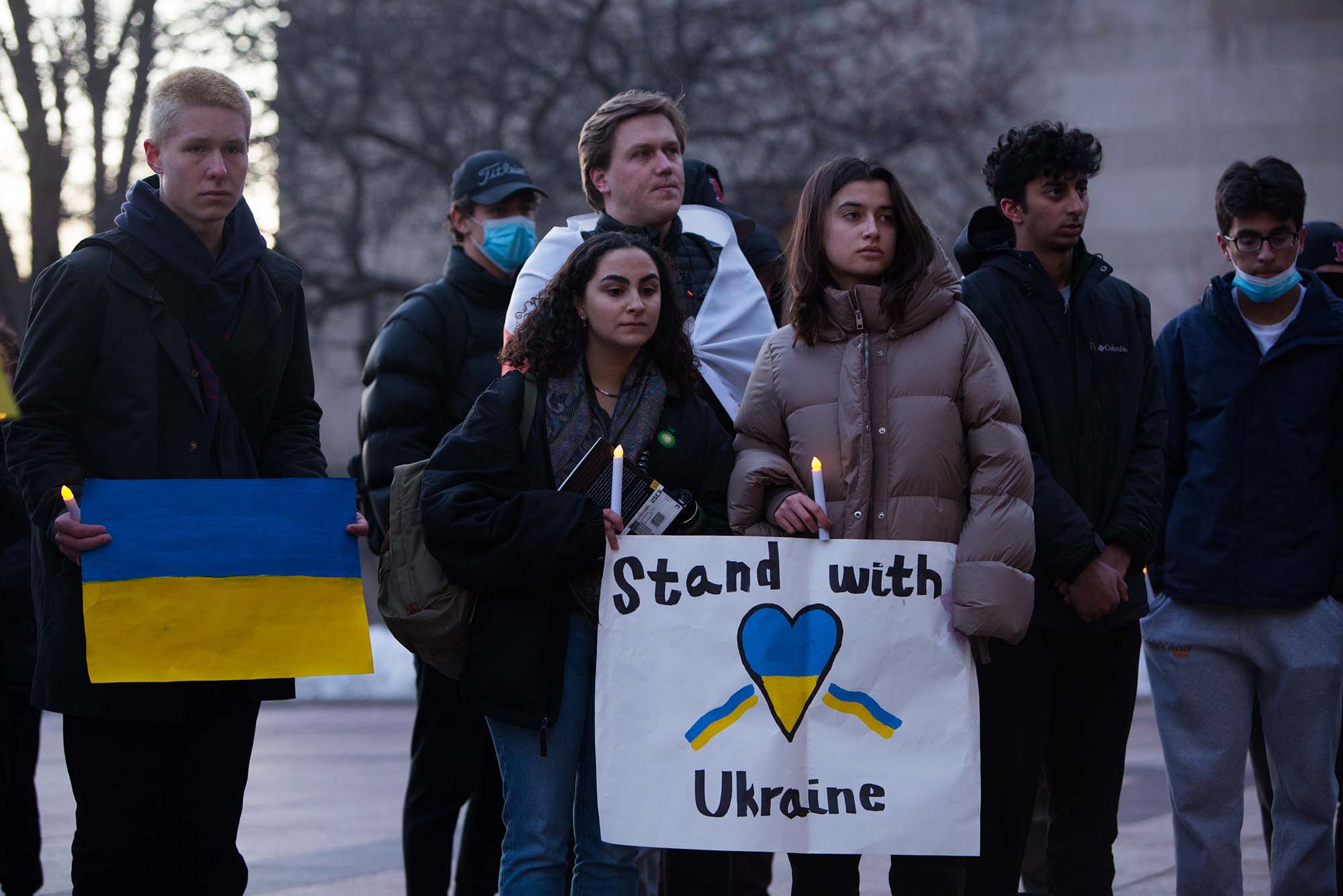 Photo of from left to right, Elvis Friberg (Questrom’24), Regina Bravo (CAS’24) and Antonia Robay (CAS’24) gathered on Marsh Plaza Thursday, March 3, 2022 for a protest and vigil in solidarity with victims of the conflict in Ukraine. At center, two students hold plastic battery candles and a sign with a blue and yellow heart that says "Stand with Ukraine"