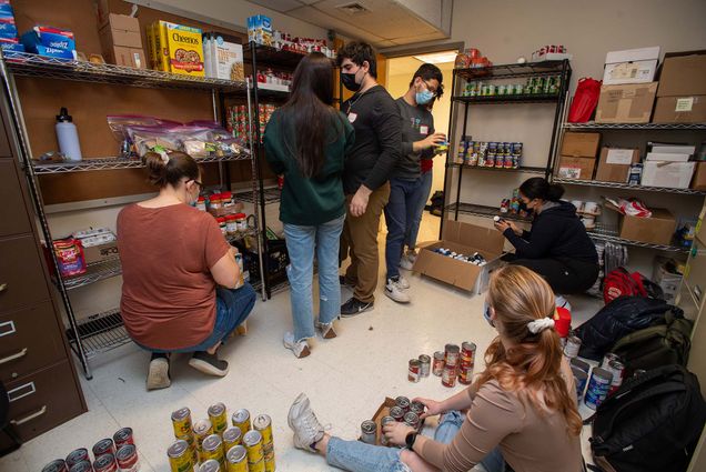 Photo of a group of masked Innovate@BU students organizing the Wheelock Food Party March 7. There are large utility racks filed with cans. some students stand while others sort cans on the ground.