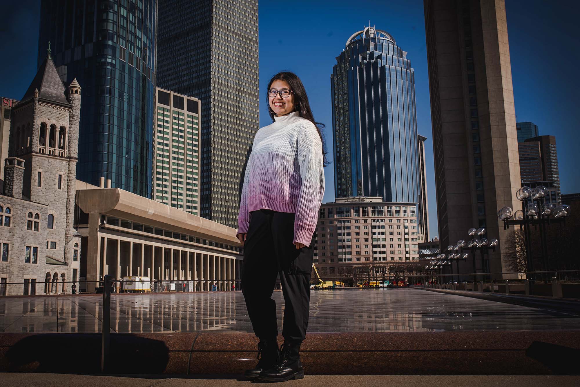 Photo of student Mira Dhakal, standing at the large reflection pool near the Christian Science Center. She smiles, wears a white and pink gradient sweater, and has long brown hair and glasses. The Boston city skyline and blue sky are seen behind her.