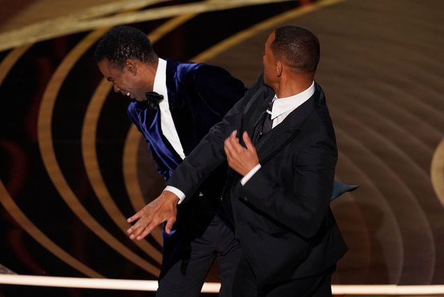 Photo of Will Smith, right, slapping presenter Chris Rock on stage before presenting the award for best documentary feature at the Oscars on Sunday, March 27, 2022, at the Dolby Theatre in Los Angeles. Both men are dressed in suits. Rock winces in pain and shock, and Smith's hand is at the end of its swing.