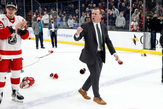Photo of Boston University Men's Hockey head coach Albie O'Connell raising his right first in the air after the Beanpot final between the Boston University Terriers and the Northeastern University Huskies on February 14, 2022, at TD Garden in Boston, Massachusetts. He is a white man with short hair and wears a suit and formal shoes, and appears to be cheering. BU Hockey players are also seen cheering and wearing championship hats.