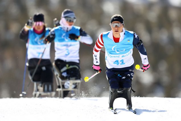 Photo of Oksana Masters, a white woman and double amputee, on Team USA who skis on a black pair of adaptive skis during an Official Training Session at Zhangjiakou National Biathlon Centre on March 03, 2022 in Beijing, China. She wears sunglasses, looks very concentrated, and two other paralympians are seen skiing behind her.