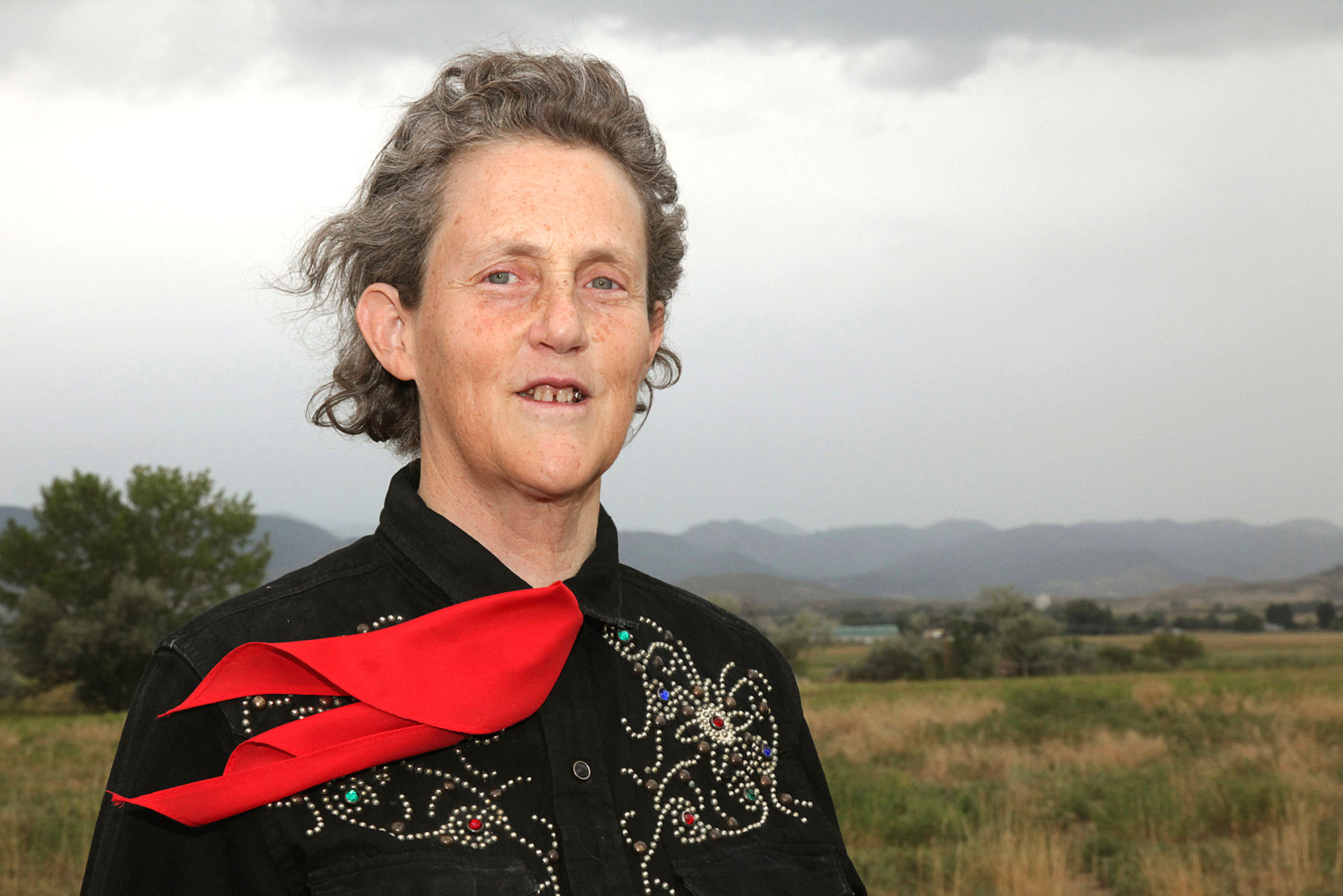 Photo of Temple Grandin. She slight smiles, has short gray hair that curls up, blue eyes and freckles. She wears a bejeweled black button up and a red neck tie. Behind her, a field and small hills are seen on an overcast day.