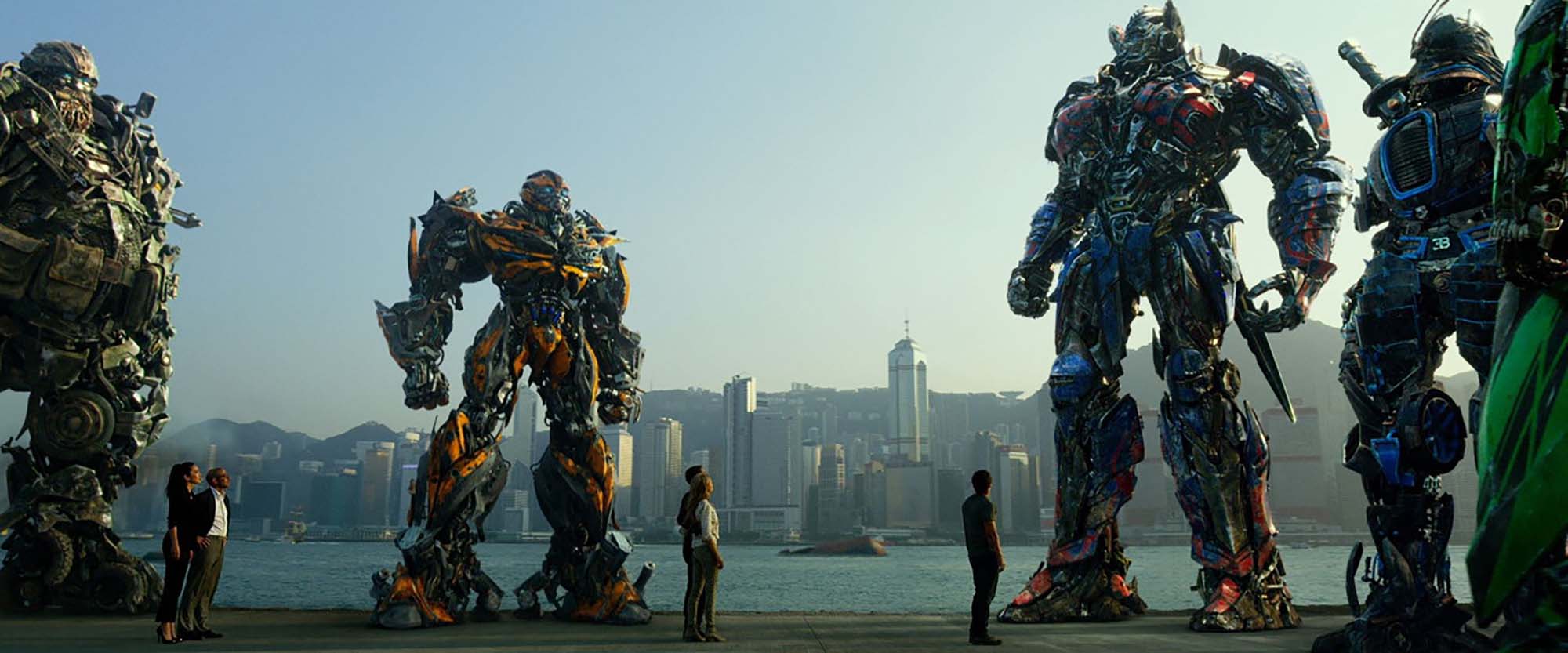 Still taken from the movie "Transformers: Age of Extinction" (2014). In the still, four larger transformers stand around a group of five humans, the movie's protagonists, who look up at them in awe. In the background, the Hong Kong city skyline is seen.