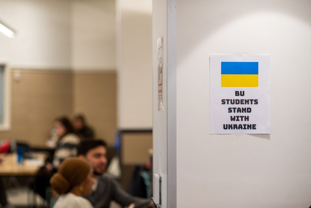 Photo taken in the GSU dining area. On a white wall, a piece of paper with a Ukrainian flag and text that reads "BU Students stand with Ukraine" hangs on the wall. In the background, blurred, students eat.