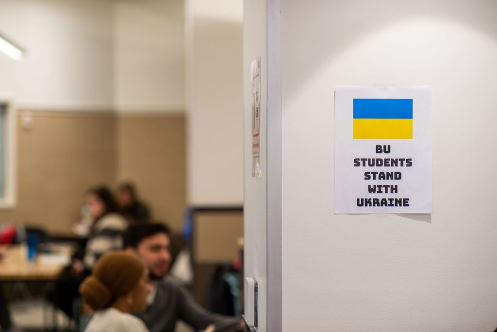 Photo taken in the GSU dining area. On a white wall, a piece of paper with a Ukrainian flag and text that reads "BU Students stand with Ukraine" hangs on the wall. In the background, blurred, students eat.