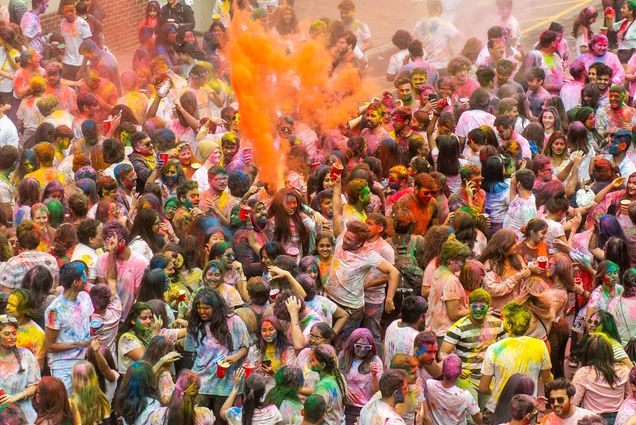 Aerial photo of BU and non-BU students celebrating Holi on Cummington Mall Rd on March 20 in-person for the first time in three years. Most young people in the crowd wear white or light colored shirts, and a cloud of orange dust rises in the air. The people are covered with an array of bright colors and look joyful as they talk with their friends.