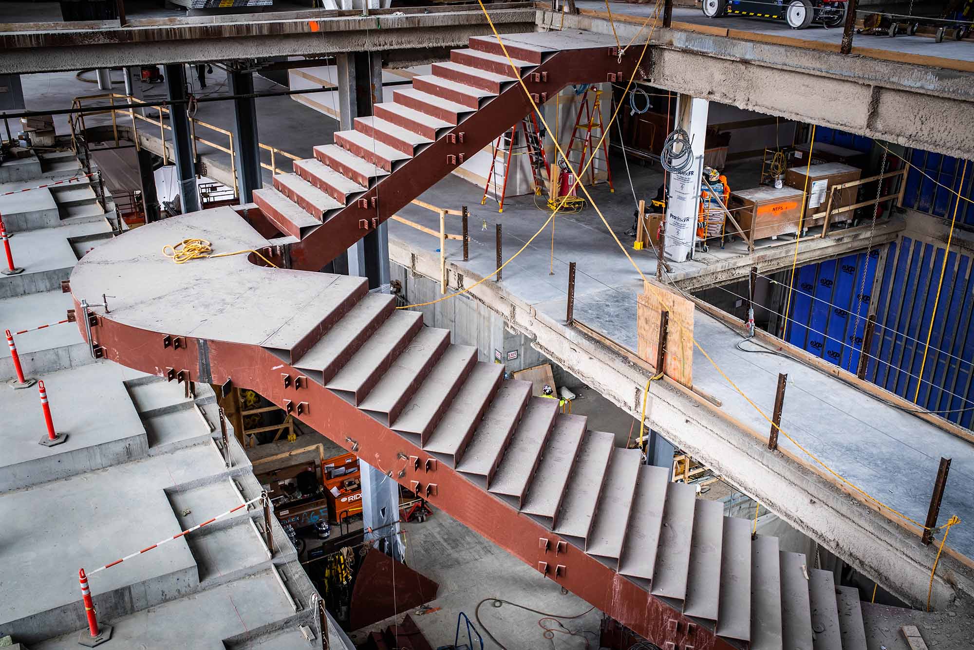 Photo of the butterfly staircase being installed in the new data sciences building. Two large staircase connect at a curved center, the sides of the staircase are maroon and constructed from large steel beams. Above and below the staircase, the construction equipment and the skeleton of the building under construction are seen.