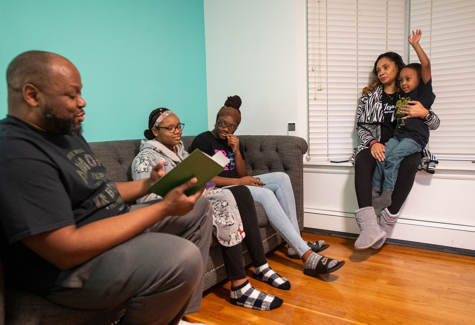 Photo of the Davis family. A Black man reads the bible to his wife and three children, two of which are seated on a sofa beside him