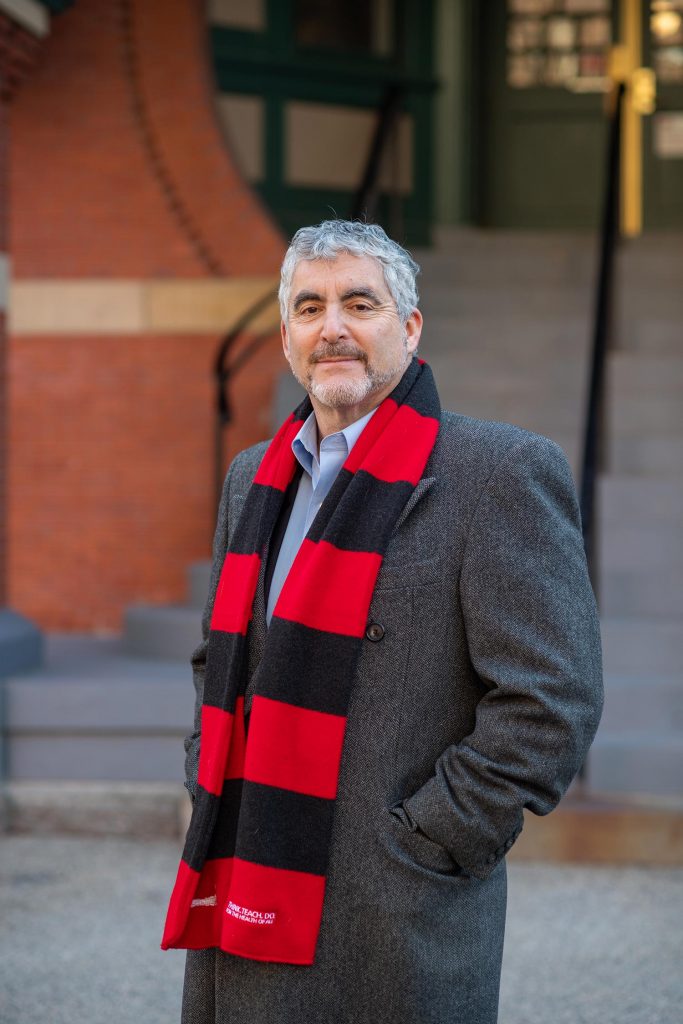 Photo of Craig Ross (SPH’14) in a long, gray winter coat and a black and red scarf standing outside a brick building.
