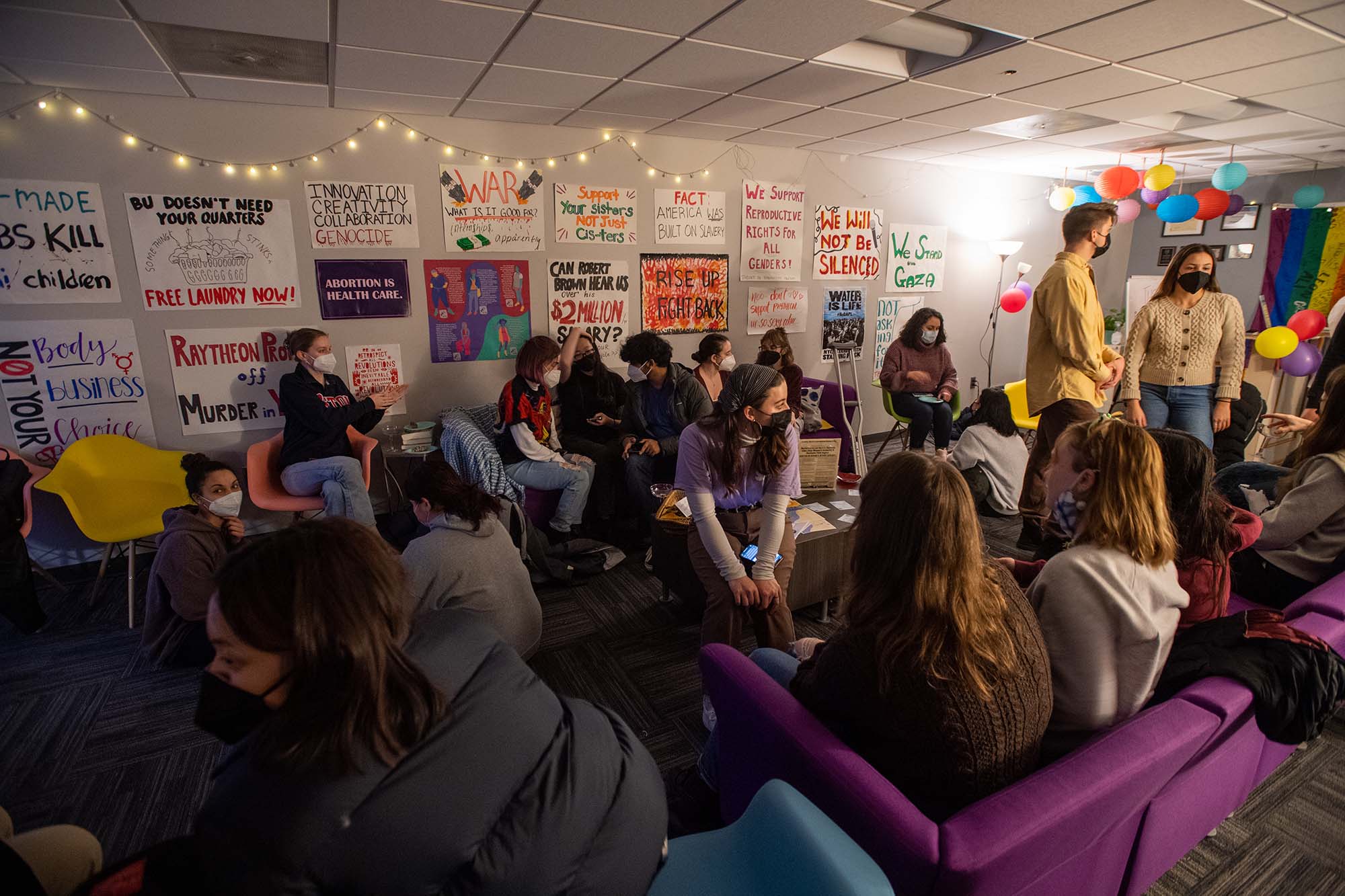 Photo of masked SRF members and supporters gathering in the Center for Gender, Sexuality & Activism during the vending machine launch party. Students are seen sitting on couches and tables and standing as they congregate in a room decorated with various handmade poster and string lights.