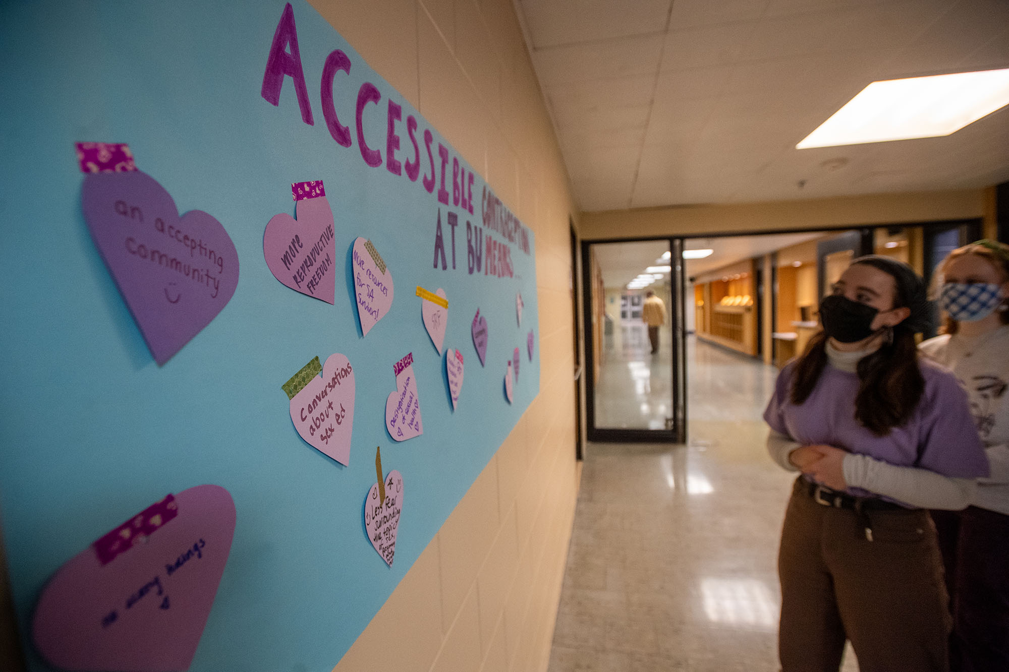 Photo of a wall set up by Students for Reproductive Freedom for students to share thoughts. A masked, female student looks on at a large oversized sheet of blue paper with various pink and purple hearts filled with writing on them.