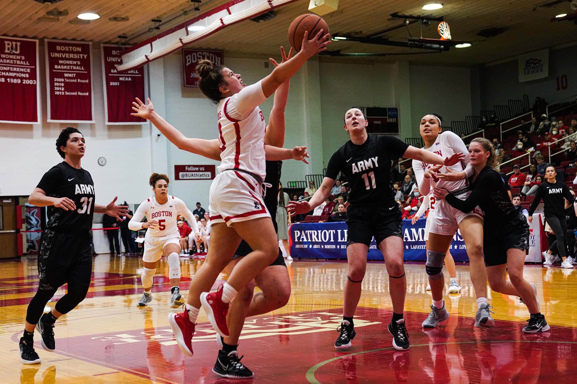 Photo of BU women's basketball Guard Alex Giannaros (CAS 25) making a layup under Army's tight defense during the PL quarterfinal game at Case Gym. A tall women wearing a white basketball uniform is mid-layup as various player in black and red uniforms react around her.
