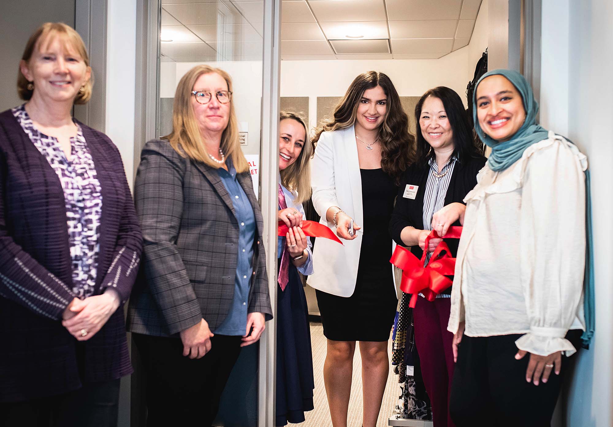 Photo of a smiling group of woman, all in business causal attire, smiling in front of a cut red ribbon