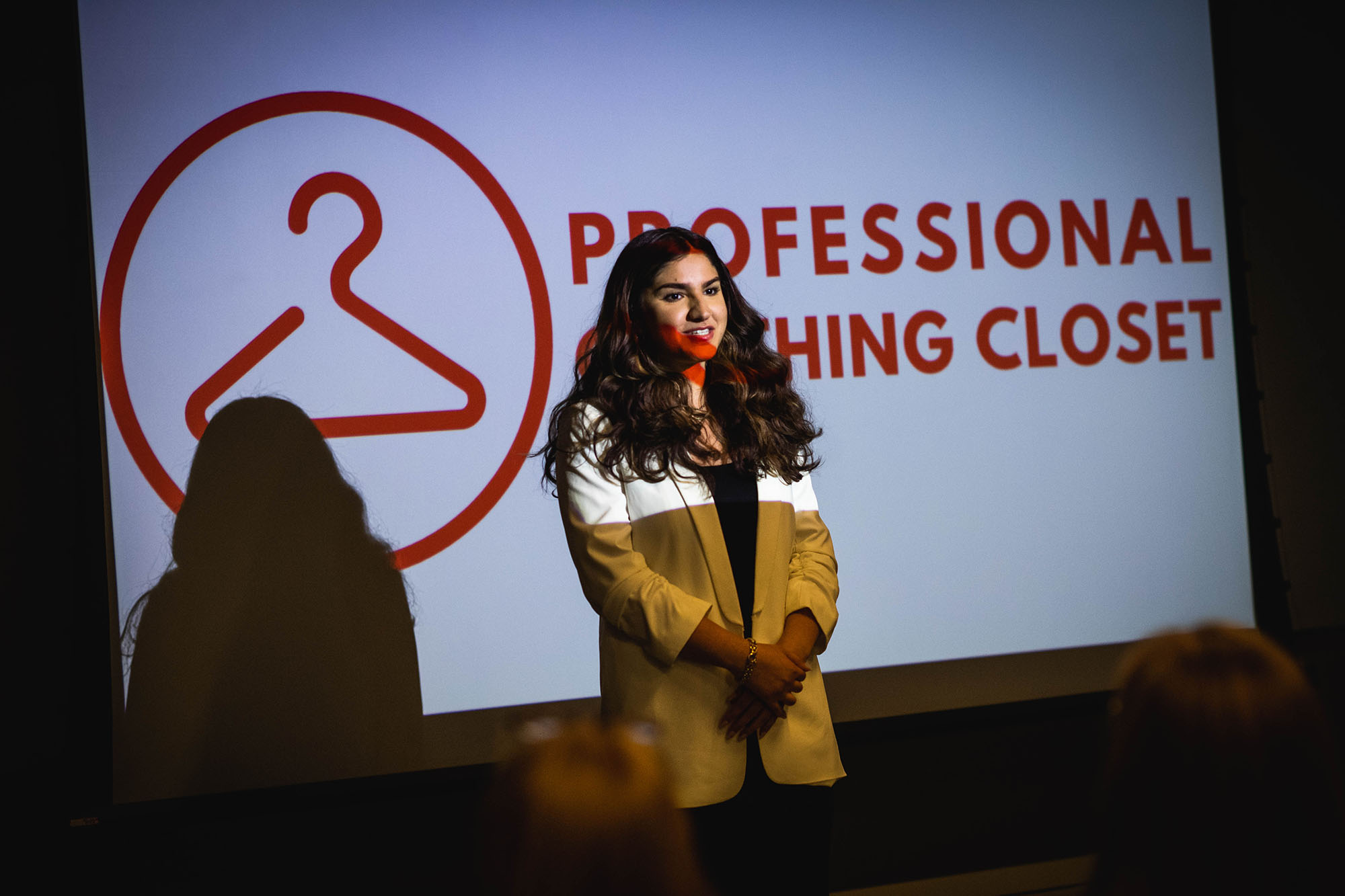 Photo of Tima Dasouki speaking at the opening of the new Professional Clothing Closet. A young, brown woman wearing a white blazer and black dress speaks in front of a project screen that reads "Professional Clothing Closet"