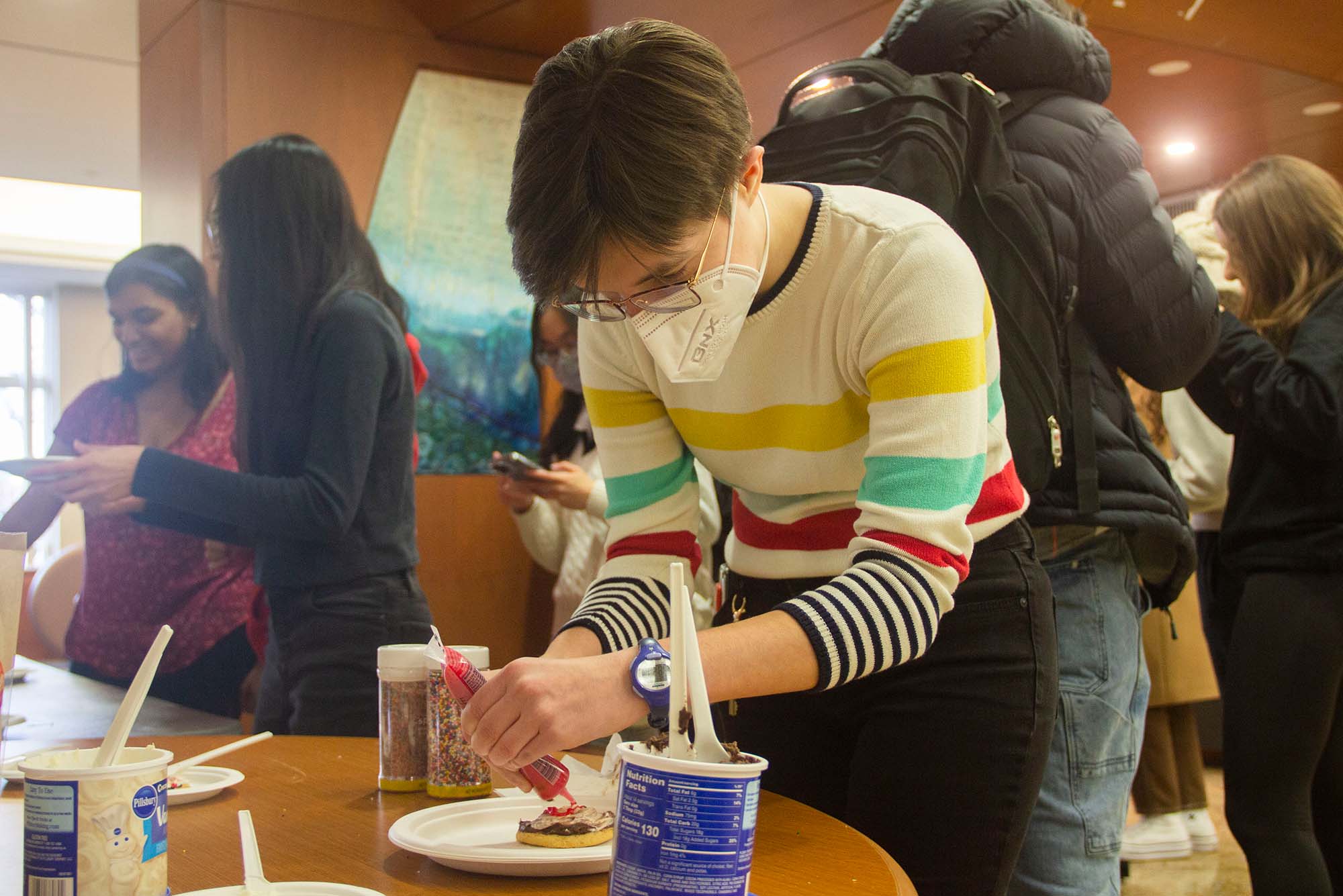 Photo of Kathryn Larkin (CAS’25), wearing a white sweater with colored strips and a face mask, looking intently at a cookie they're adding red frosting too. Containers of vanilla and chocolate frosting are seen in the foreground and other students milling about are in the background.