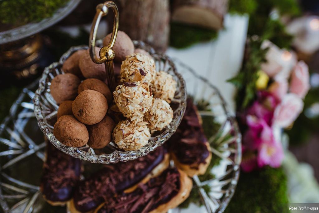 Photo of Petite eclairs and assorted chocolate truffles. An afternoon tea tray features truffles on the top tier and eclairs on the bottom. The blurry background features pink and white flowers.