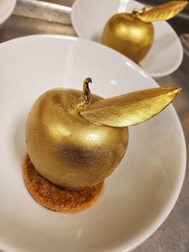 Photo of The Golden Apple, which features cinnamon diplomat cream, fresh apple and brandy liqueur jam, a white chocolate shell, and gold luster dust. A golden apple sits in a shallow, white bowl