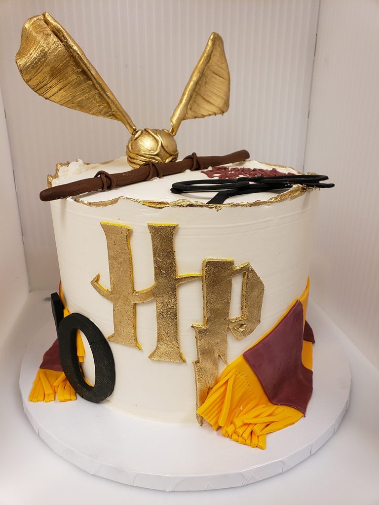 Photo of a Harry Potter birthday cake. A white cake features a large gold "HP" logo, Griffindor scarf, and golden snitch and wand as toppers.