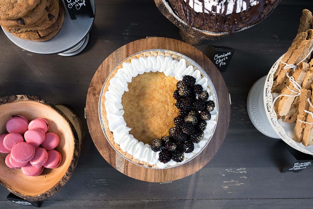 Photo of a buttermilk pie. it is served with whipped cream and fresh blackberries and is surrounded on a black table by other various desserts such as macarons and pie.