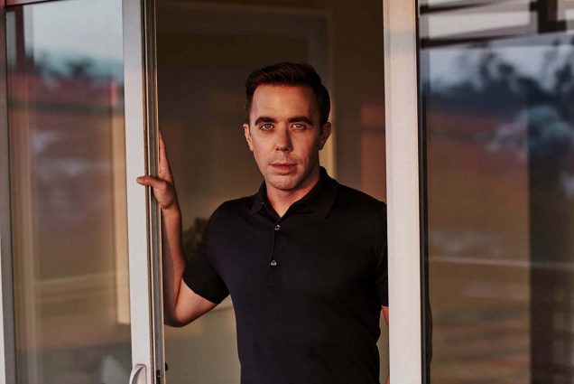 Photo of Erich Schwartzel (COM’09), a light skinned man with short brown hair. He wears a black polo shirt and leans on a sliding glass door with this right hand.