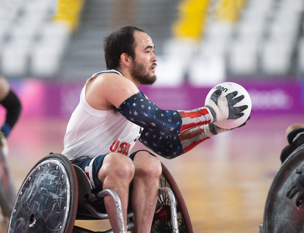 Photo of Wheelchair rugby player Chuck Aoki catching or about to pass the ball. He is sweaty and wears sleeves with American flags on them. 