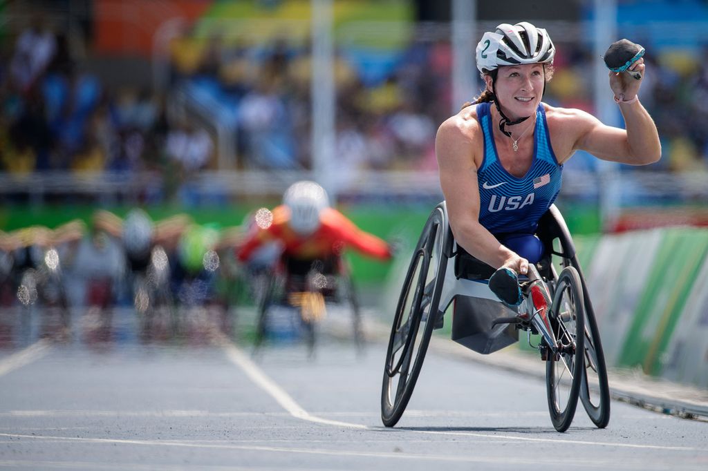 Photo of Track and field star Tatyana McFadden, who is a dominant force in women’s wheelchair racing. She raises her first in the air and smiles; she wears a helmet, and blue jersey, as she zooms across the finish line.