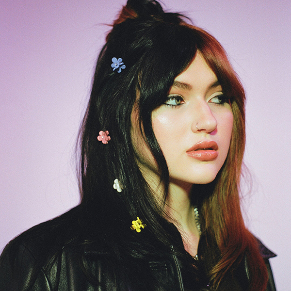 Photo of GAYLE, a white woman with black hair and dyed brown bangs, looks to the right with a curious expression.  Small flower clips and sprinkles in her hair as she stands in front of a muted pinkish purple backdrop