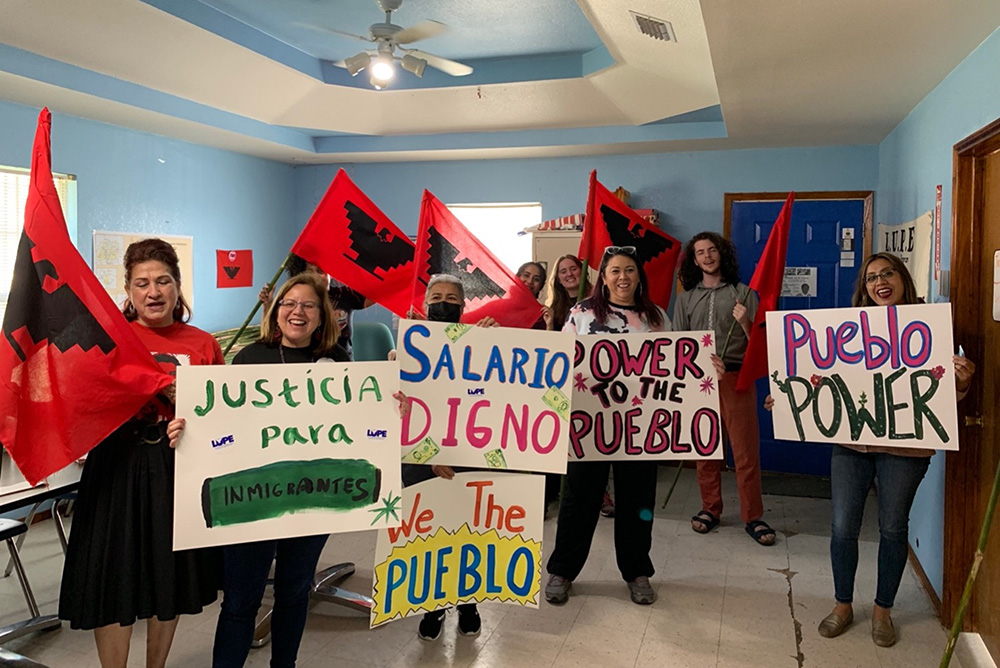 Photo of a group of people in a light blue room holding red flags and white signs with Spanish writing up