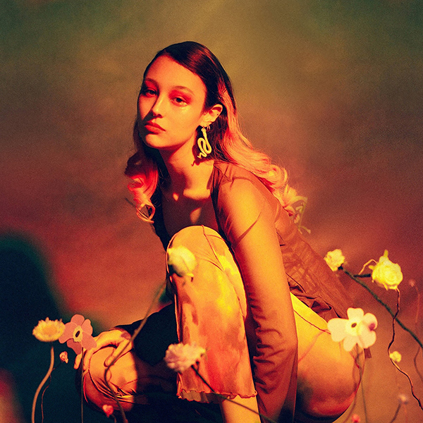 Luna Li album cover "Duality". The image shows Luna Li, squatting in front of a background with various dried flowers in the foreground. The entire scene and image are tinted with dark oranges and greens, giving off a soft tone.