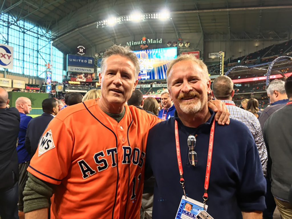Photo of Jerry Crasnick with a press pass on at the 2017 World Series in Houston with BU grad and former 76ers coach Brett Brown, who wears an orange astros jersey and has his arm around Crasnick.