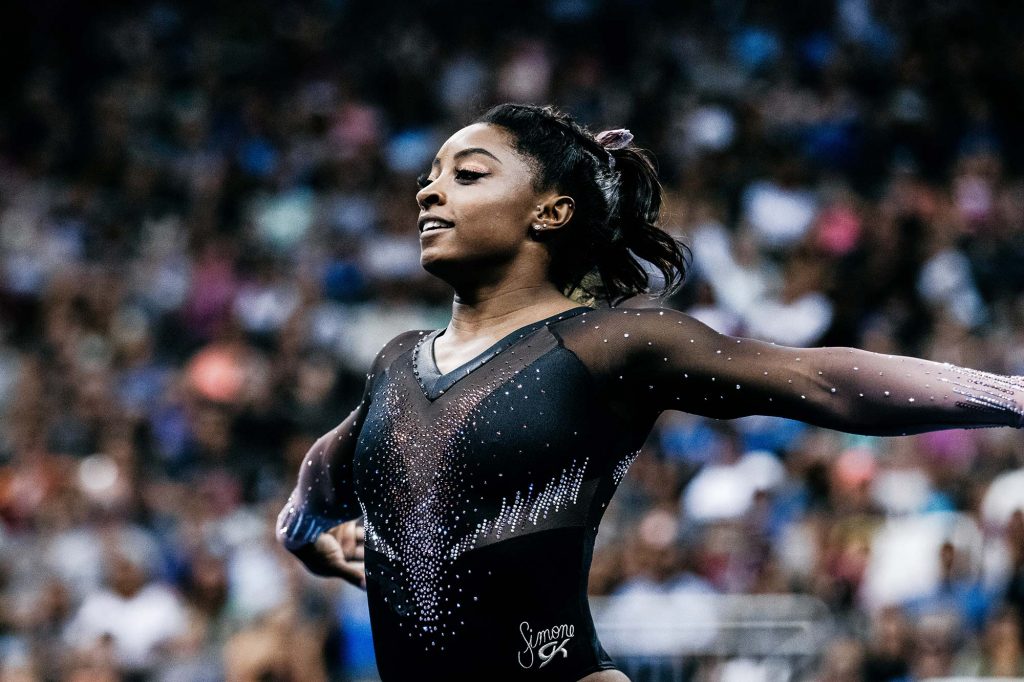 Photo of Simone Biles in a black, sparkly leotard with her arms extended back as if she has just finished a routine. A blurred crowd of people are seen behind her.