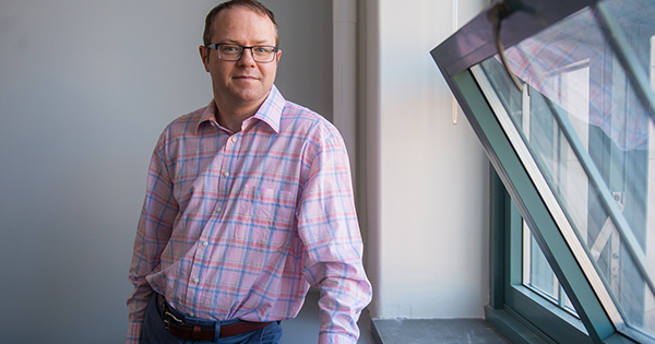 Photo of Benjamin Sovacool. A middle-aged white man wearing glasses and a light pink plaid shirt and jeans poses next to an open window and lightly smiles at the camera.