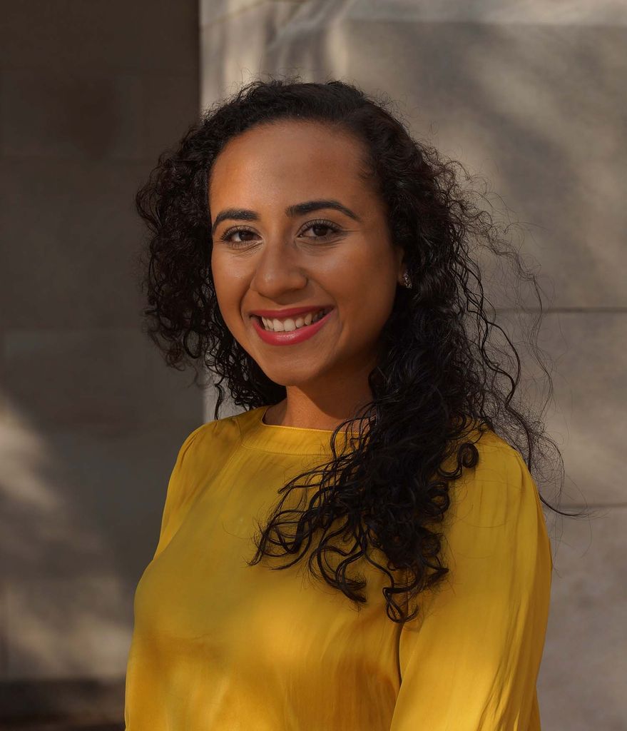 Photo of Lara Werneck, a young woman of color with long curly brown hair. She smiles, leans against a wall, and wears a yellow long sleeve.