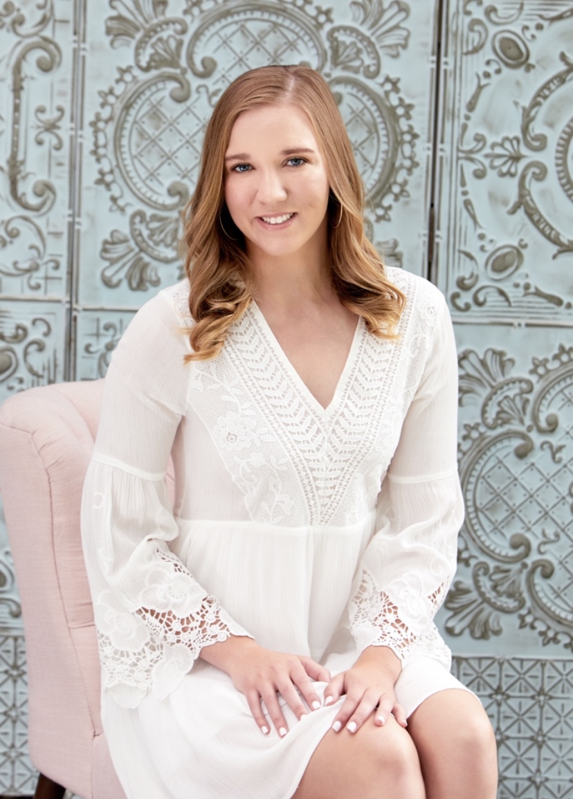 Photo of Allie Barwind, a white woman with blonde hair, who smiles and wears a white dress with lace sleeves. She sits on a pink chair in front of a washed tiled wall.
