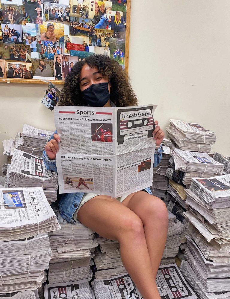 Photo of Colbi Edmonds, a brown-skinned woman with curly shoulder length hair, wears a black face mask and sits on top of a pile of printed copies of the daily free press. She opens up to the sports page and smiles from underneath her mask.