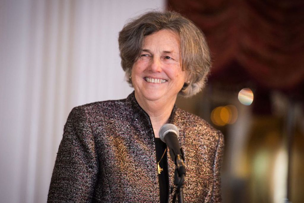 A photo of Phyllis Zagano in a brown blazer jacket with a microphone on a podium in front