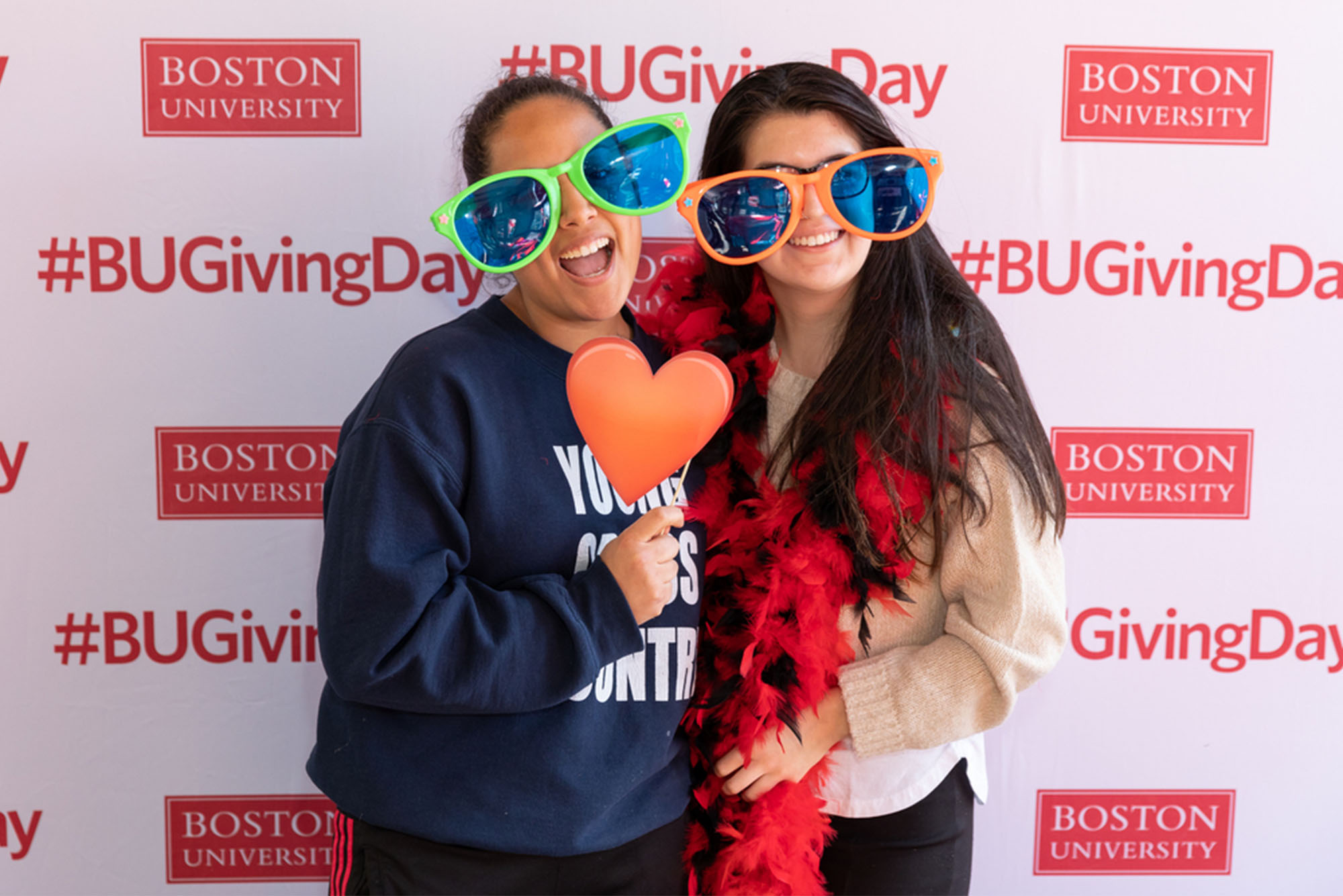 Two friends wear large sunglasses in front of a photo board with writing "#BUGivingDay" in red letters