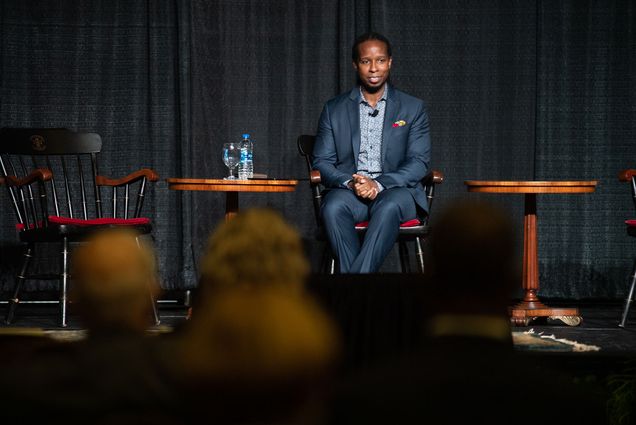 Photo of Dr. Ibram X. Kendi at What the Science Tells Us: Racial Health and Economic Inequities during the Pandemic Symposium presented by The BU Center for Antiracist Research in the Metcalf Hall October 1. He wears a blue suit and a patterned blue button down. He sits on stage with hands folded in lap as he looks on to the audience.