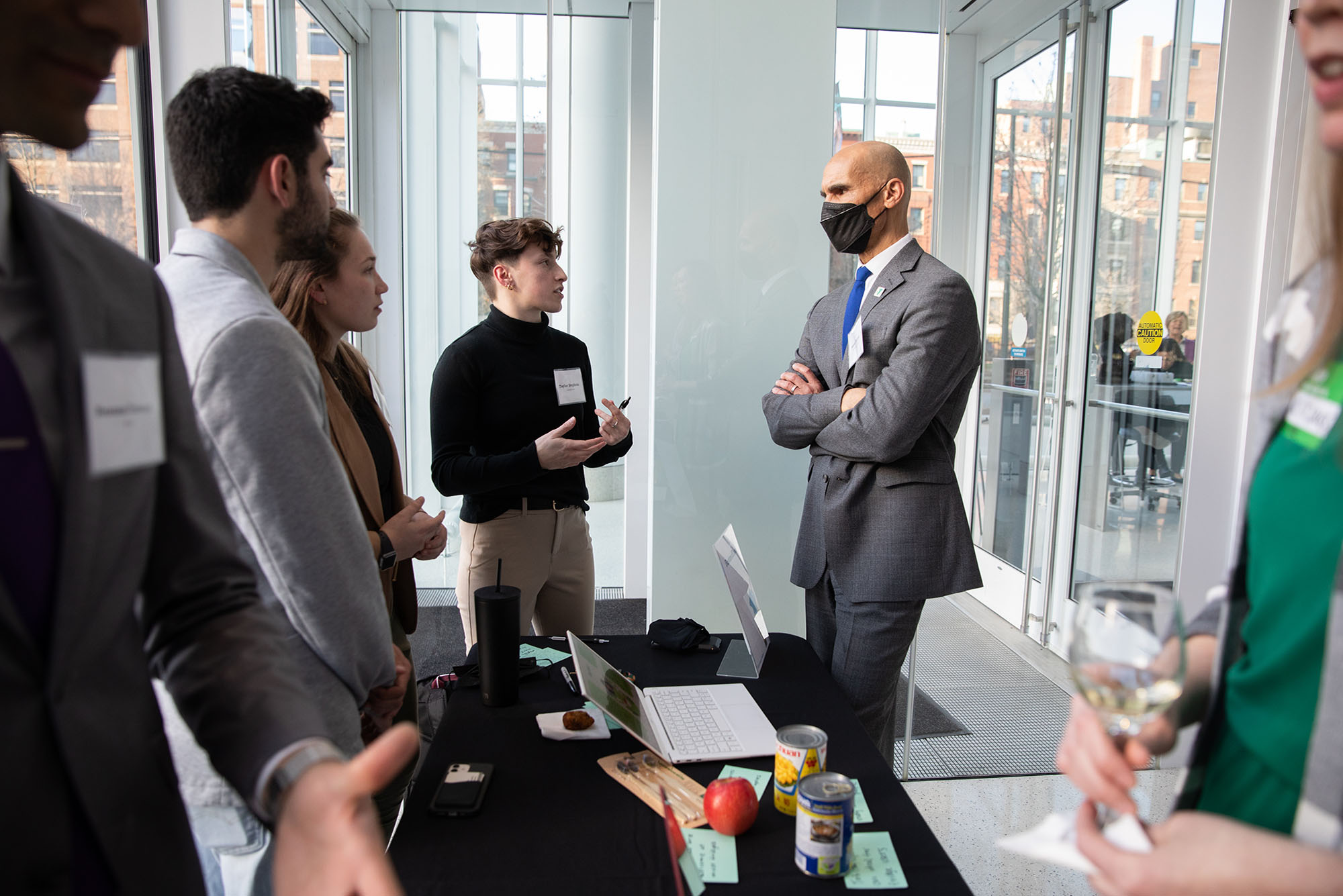 Photo of Fostered Food team members Matt Blanco, Hayes Hart-Thompson, and Taylor Stephens explaining their idea to James Morton. 3 students in business casual attire speak to a tall, tan masked man in a grey suit.