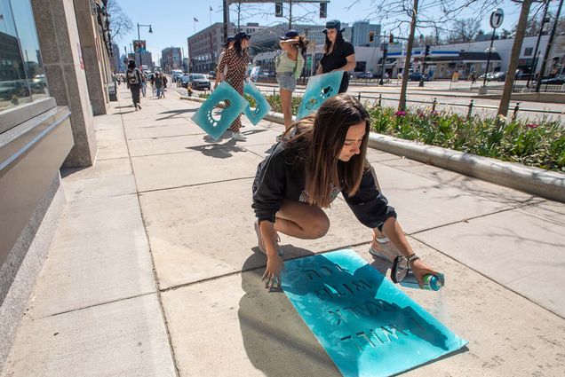 Photo of SARP student ambassador Hannah Zobair, a young white woman with long brown hair and wearing a black sweatshirt, holding a large blue stencil to a sidewalk and spraying a can of light blue paint onto it on a sunny day. Three other young women stand behind her with similar stencils as they survey the ground.