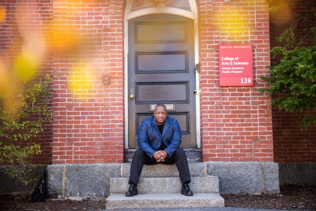 Photo of Louis Chude-Sokei, an older black man wearing a dark blur shirt and black shoes sits with hands folded in front of him on the steps of a brick building. A red sign on the building reads "Boston University College of Arts and Sciences; African American Studies Program"