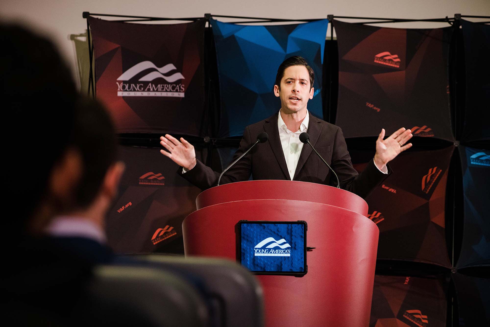 Photo of Michael Knowles during his talk on "Teaching Kids Sex and Gender Ideology," with BU YAF at the Law Auditorium on Thursday. Knowles is a younger white man with short brown hair who wears a suit and extends his arms outwards as he speakers. The podium is red, has a blue young Americans logo on it, and he stands in front of a background with the young Americans logo in blue and red.