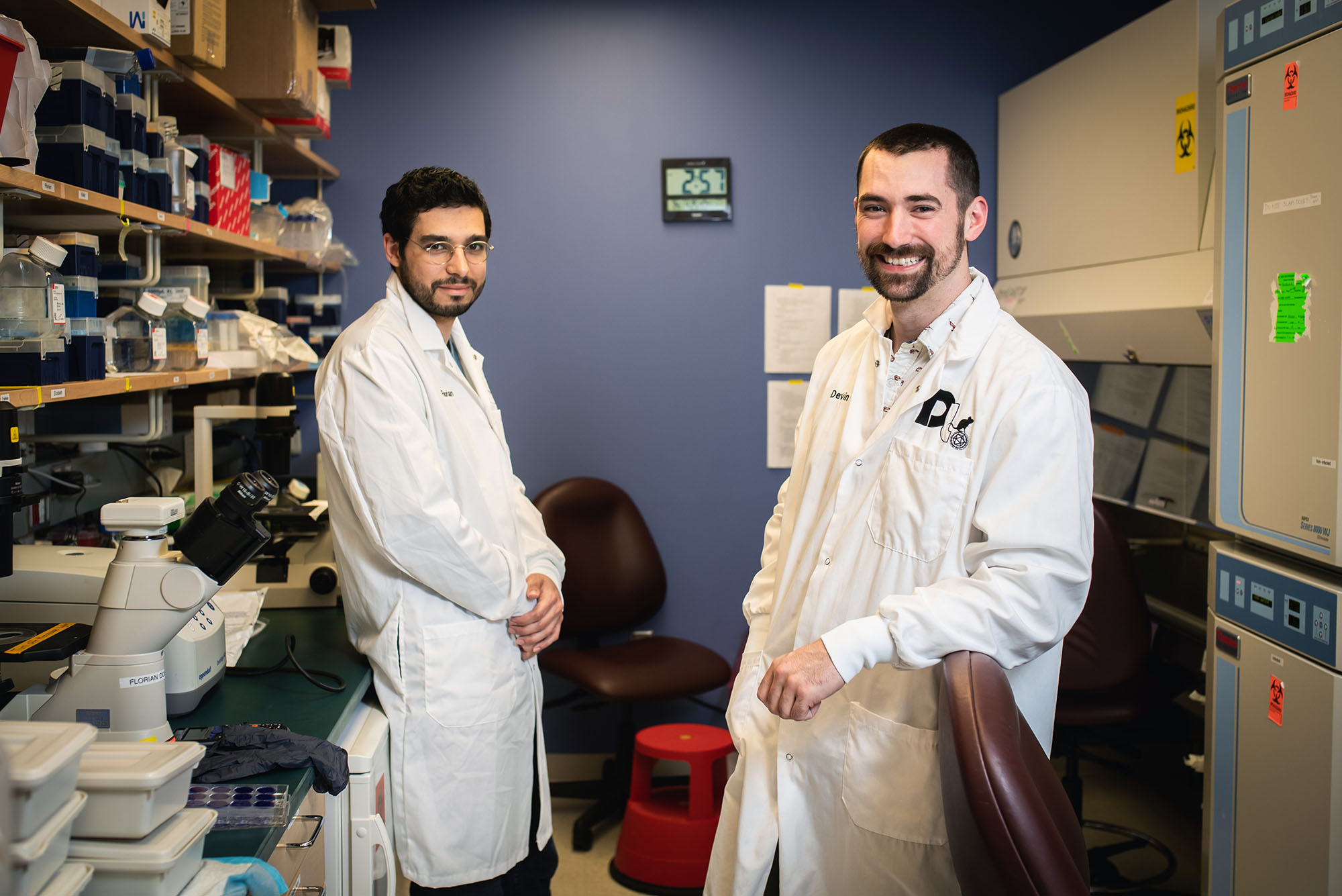 Photo of Florian Douam (left) assistant professor, microbiology, and Devin Kenney, a PhD student, posing for a photo at National Emerging Infectious Diseases Laboratories. The both wear white lab coats and stand facing each other with their bodies and heads tilted towards the camera in front of a purple-blue wall. Lab shelves can be seen on either side of them.