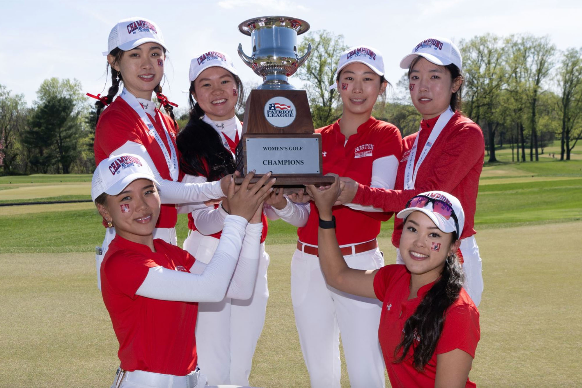 Photo of the BU Women's Golf team posing with the Patriot League trophy. From left, Kristen Min Ju Kim, Flair Kuan, Hanako Kawasaki, Alice Fan, Christy Chen, and Victoria Takai all stand and kneel around the trophy, each using one or two arms to touch and hold it up in the center of themselves, as they hold up a large silver trophy with a large wooden base.