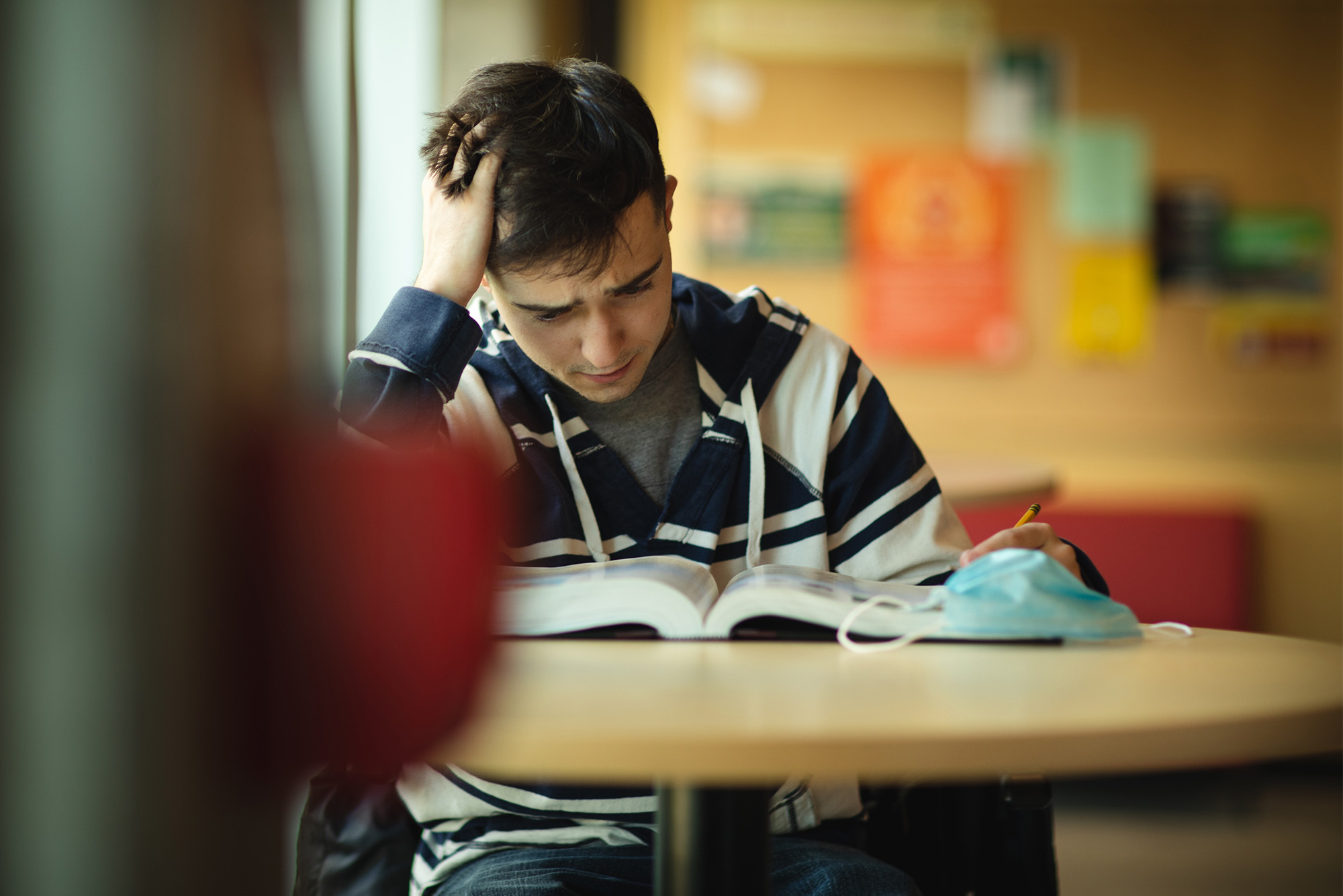 A college student appears stressed as they study at a library table with open books and a pencil