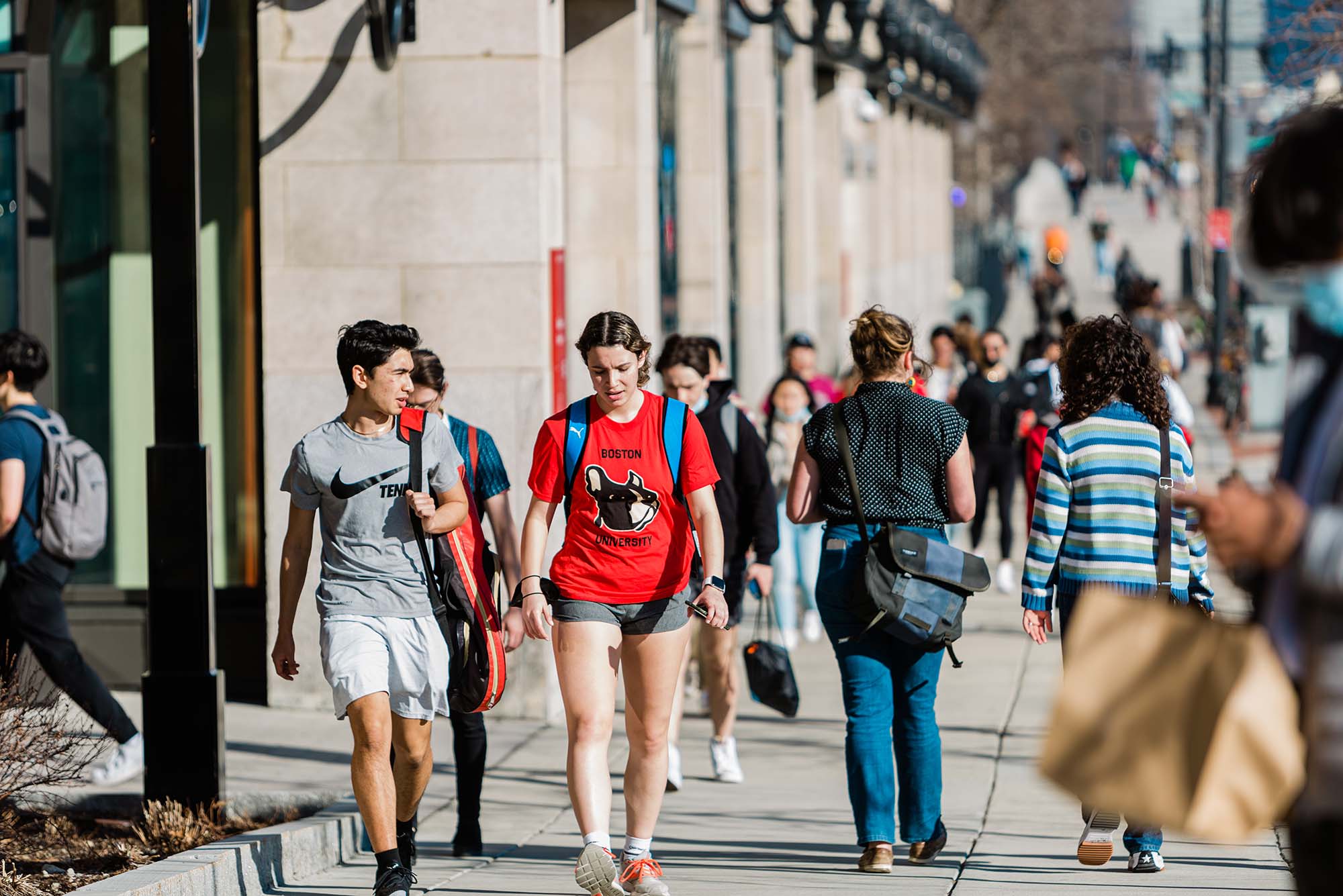 Photo of students walking down the CFA side of Comm Ave on a sunny warm day. Two students are in focus at the front left, one wears a gray nike tennis shirt, the other a red Boston University shirt.